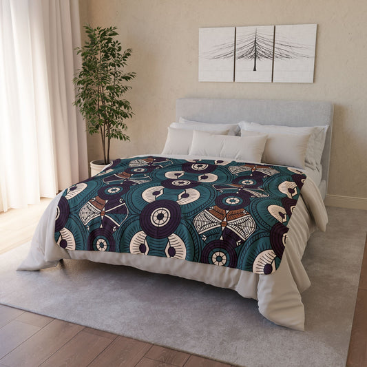 A neatly made bed with a gray headboard and white bedding, topped with a colorful, African-inspired fleece blanket in teal, navy, brown, and cream. A potted plant stands to the left and abstract artwork hangs above as light streams through white curtains.