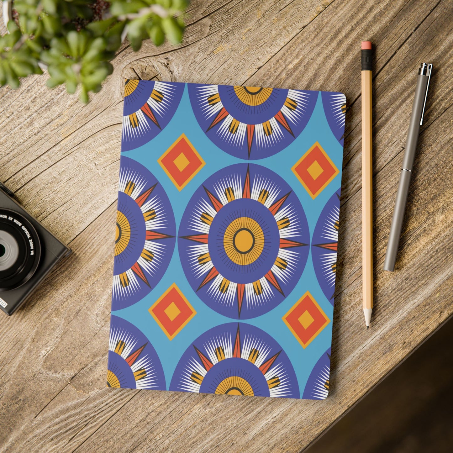 A vibrant African-inspired journal with a bold, colorful geometric pattern lies on a rustic wooden table. Nearby are a pencil, silver pen, part of a camera, and a green potted plant—perfect for daily journaling or creative notes.