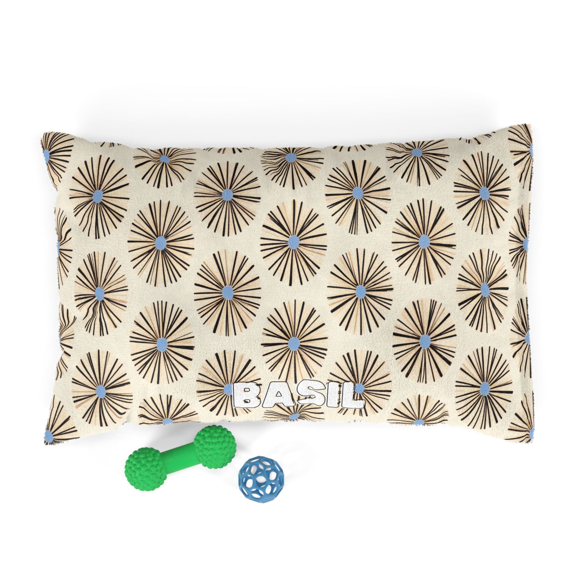 A rectangular pillow with a beige background and African-inspired brown starburst patterns with blue centers. The word BASIL is printed in large white letters at the bottom center, with two green and one blue dog toys placed beside it.
