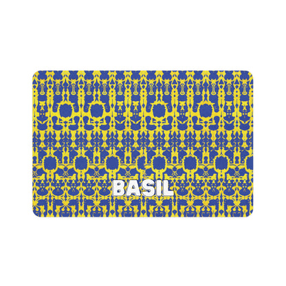 A rectangular mat with a bold blue and yellow symmetrical geometric pattern. The African-inspired, repeated shapes create an abstract, kaleidoscopic effect. The word BASIL in large, white, bold letters is centered near the bottom of the mat.