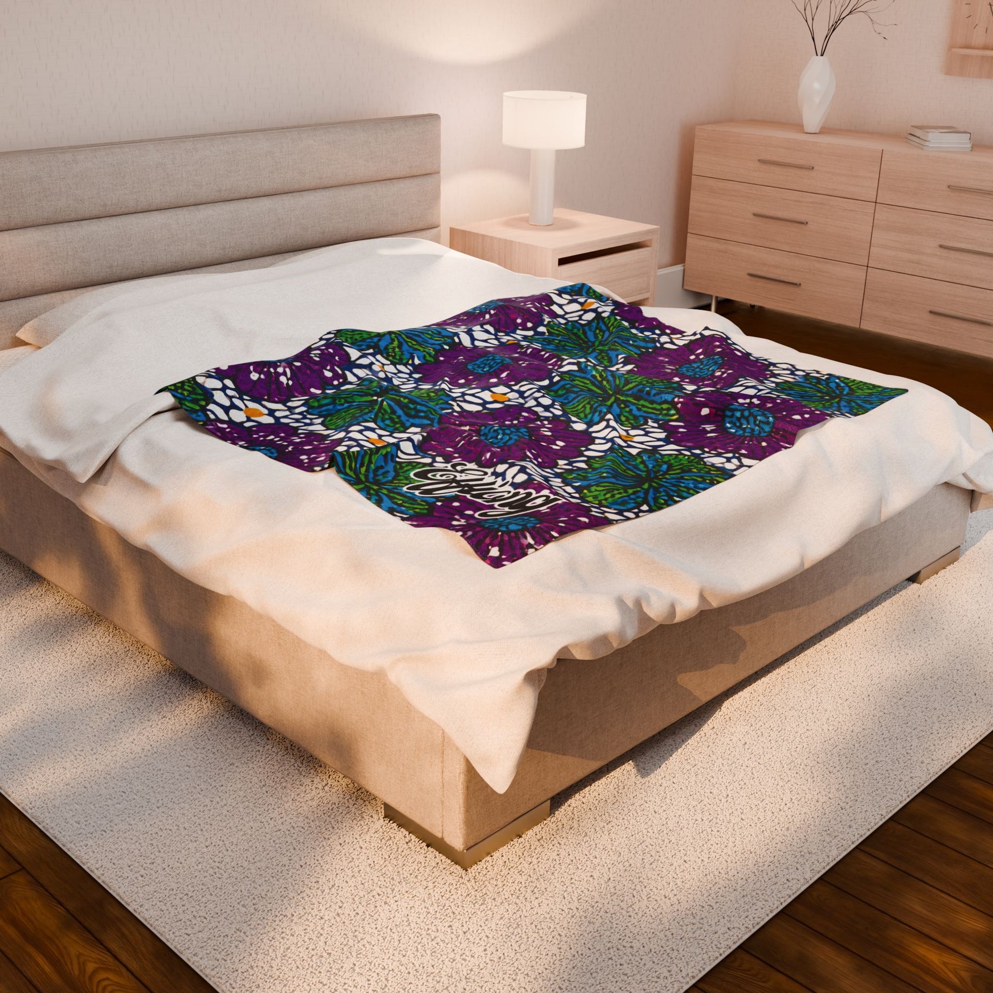 A modern bedroom with a beige upholstered bed, white bedding, and a vibrant African-inspired personalized blanket featuring green, blue, white, and purple leaf designs. The room includes a nightstand with a lamp, dresser, rug, and wood flooring.