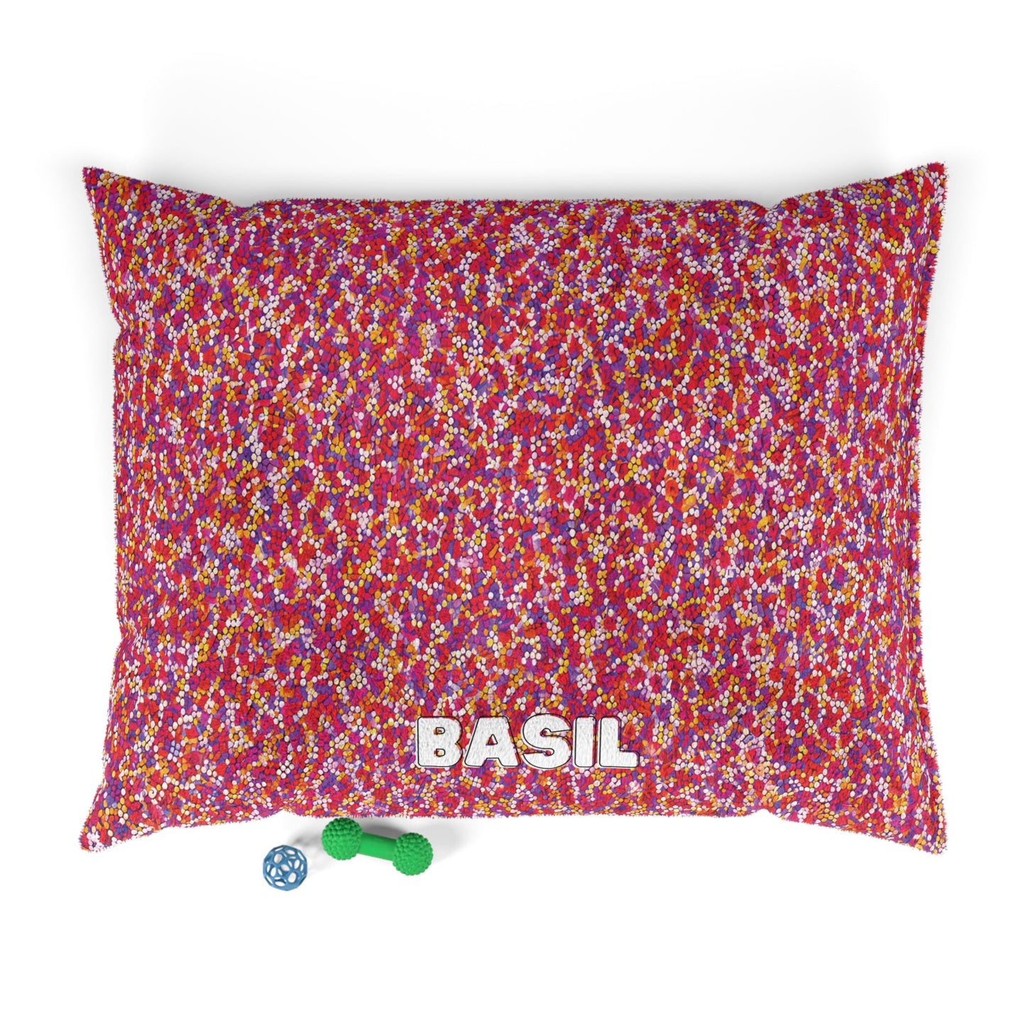 A rectangular dog bed with luxurious comfort and a red, pink, white, and yellow speckled pattern features “BASIL” in white on the front. Below are a green bone-shaped toy and blue ball with holes. The background is white.