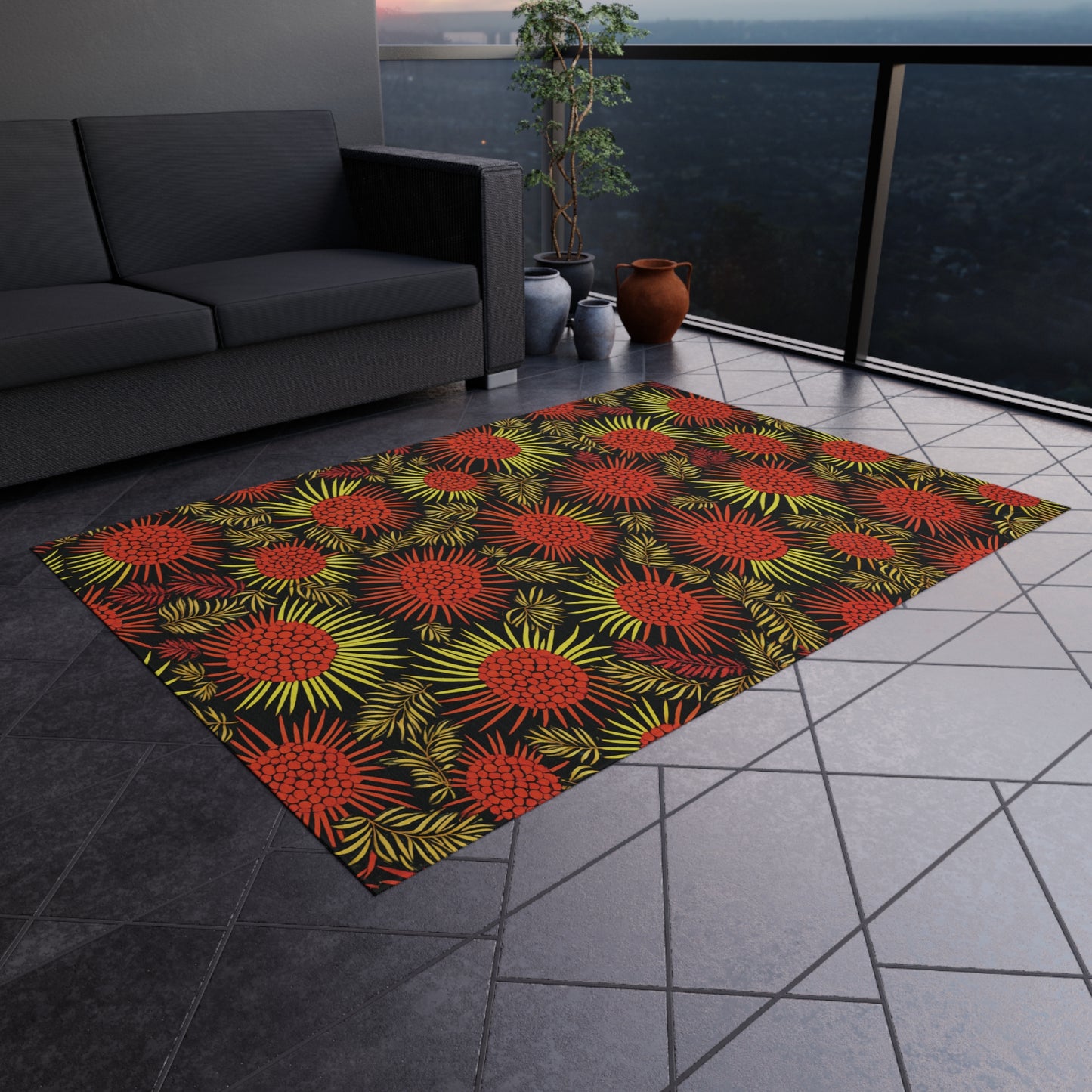 A balcony with a dark gray sofa, potted plants, vases, and an African-inspired patterned rug. The rug features a black background with red and orange floral designs and yellow-green leaves. Gray tile flooring and a glass railing overlook the city below.