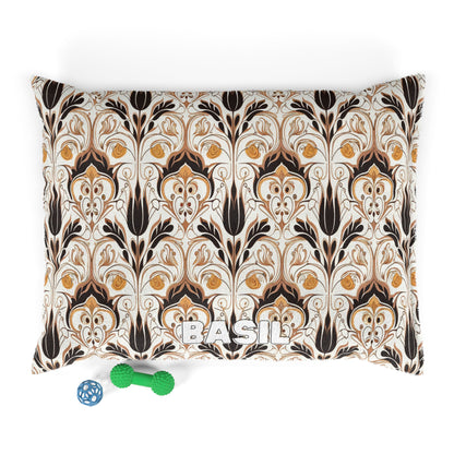 A rectangular pillow with an African-inspired, art nouveau-style floral pattern in brown, gold, and black on a cream background. The name “BASIL” is printed in white at the bottom. Two dog toys—a blue ball and a green bone—are placed below.
