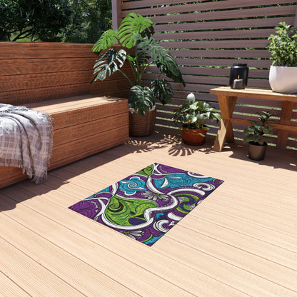 Outdoor patio with wooden flooring, a wicker bench with a gray plaid blanket, potted green plants, and a small wooden table topped with a black pitcher. An African-inspired outdoor rug featuring purple, green, and blue swirls adds vibrant flair to the space.