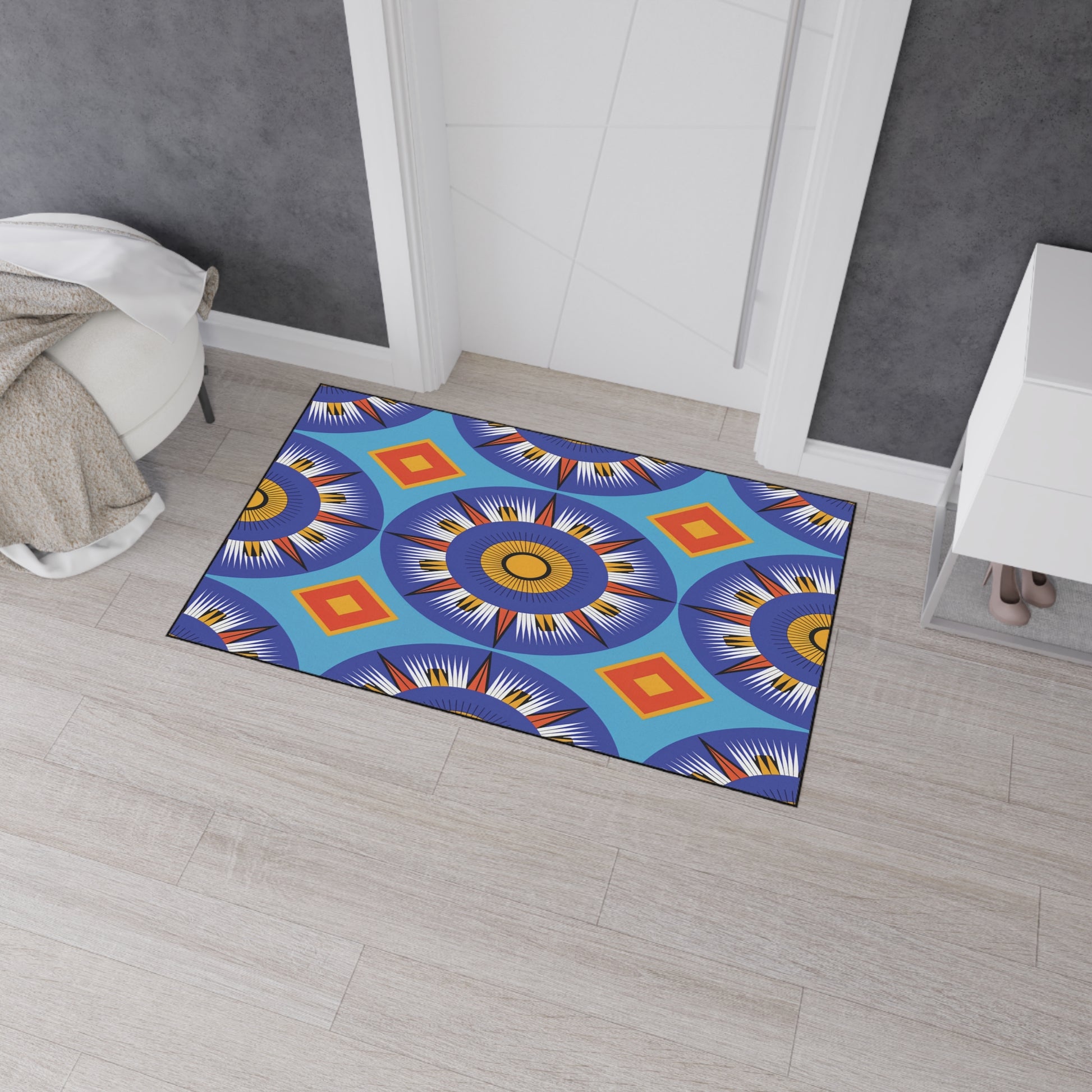 A colorful, African-inspired doormat with bold, circular geometric patterns in blue, orange, yellow, and white lies on a light wood floor near a white door. Gray walls, a white cabinet, and a beige textured throw complete the scene.