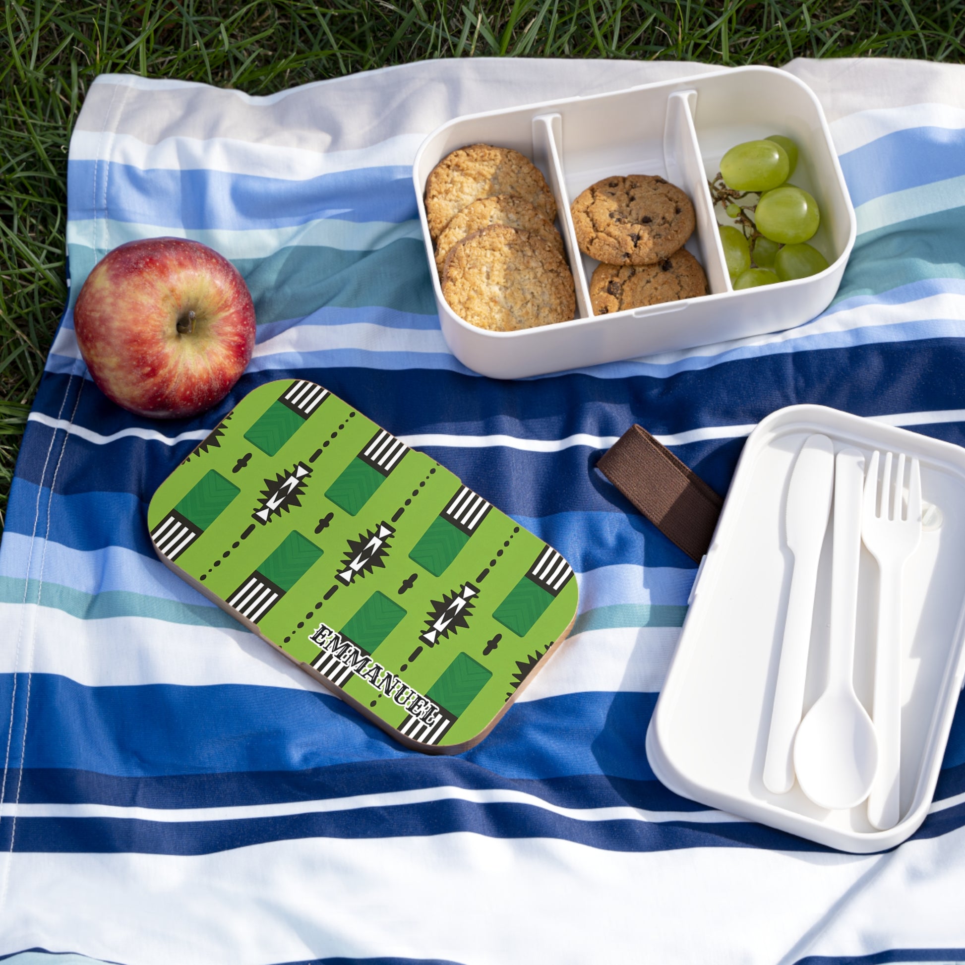 A striped picnic blanket on grass holds an apple, a green-patterned personalized lunch box labeled “EMMANUEL,” cookies and grapes in an African-inspired bento lunch box, plus a white container with a fork, spoon, and knife.