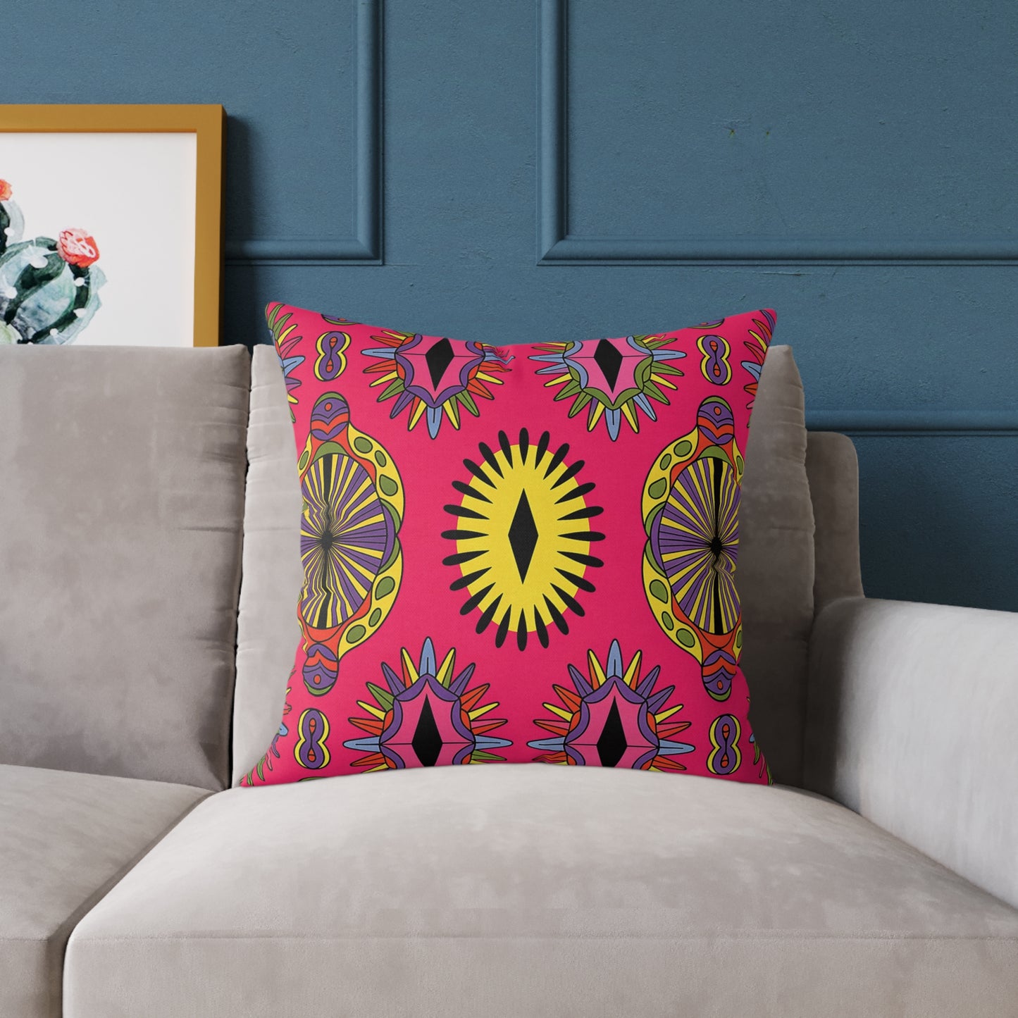 A colorful accent pillow with a vibrant geometric pattern in yellow, black, orange, and purple sits on a light gray sofa. Featuring bold, symmetrical starburst designs on a pink background, this vibrant home decor piece pops against the blue wall and cactus art.