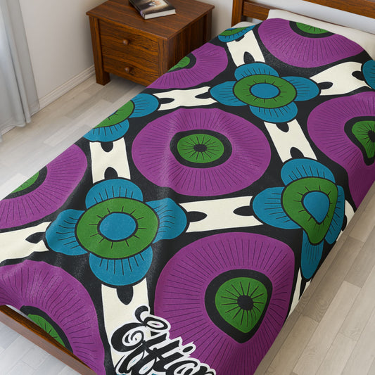 A bed features a custom velveteen blanket with bold circular patterns in purple, blue, and green on a black and white background. “Effort” is printed in script at the corner—an African-inspired personalized blanket that makes a unique gift.