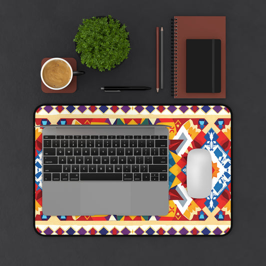 A laptop and white mouse sit on a colorful geometric-patterned Neoprene desk mat atop a dark desk. Nearby are a cup of coffee, a potted plant, writing tools, and two notebooks—one brown, one black—arranged neatly above the mat.
