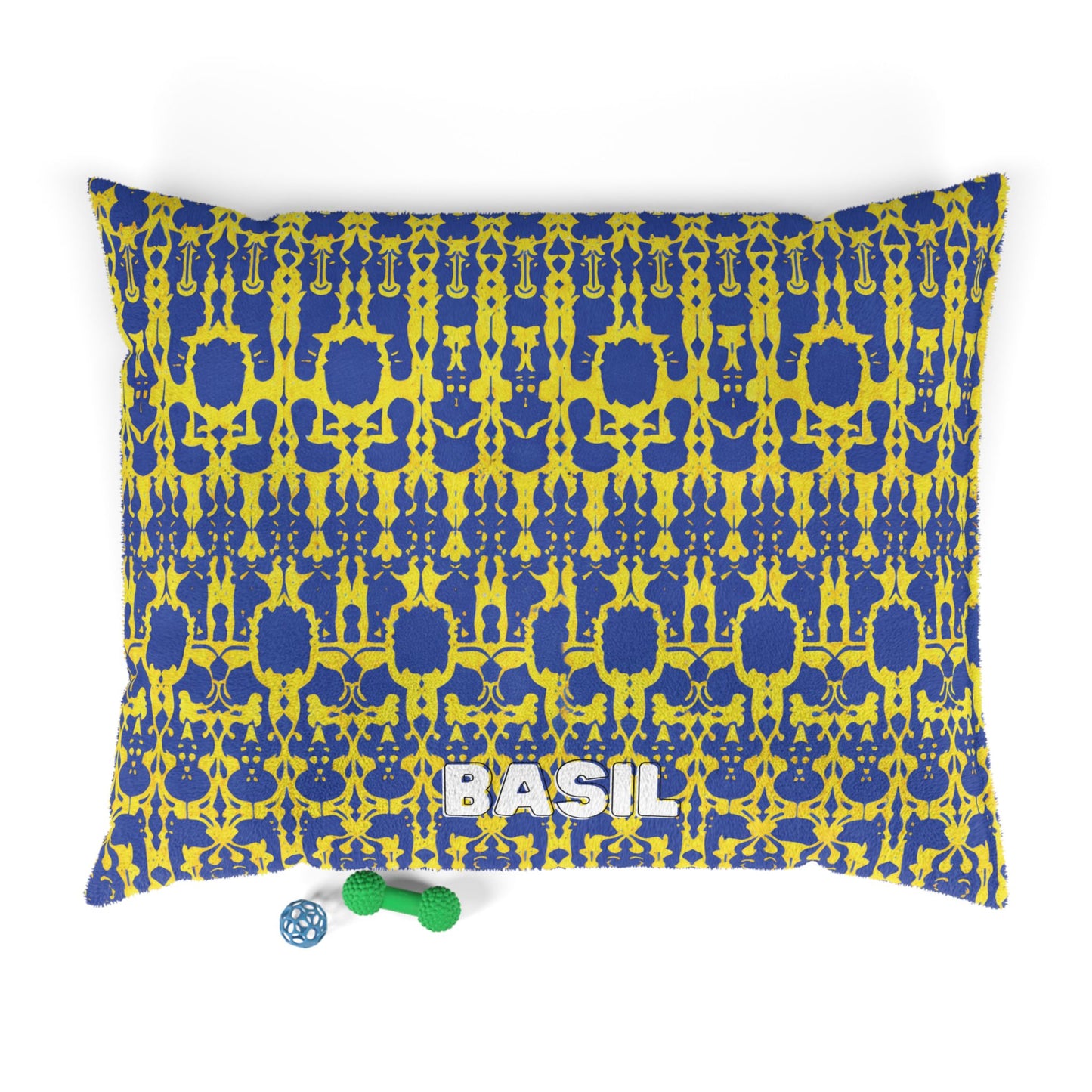 Rectangular, personalized pet bed with a blue and yellow intricate pattern offers luxurious comfort. BASIL is printed in large white letters at the bottom center. Two dog toys—a green bone and blue ball—rest on the white background nearby.