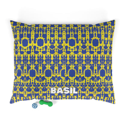Rectangular, personalized pet bed with a blue and yellow intricate pattern offers luxurious comfort. BASIL is printed in large white letters at the bottom center. Two dog toys—a green bone and blue ball—rest on the white background nearby.