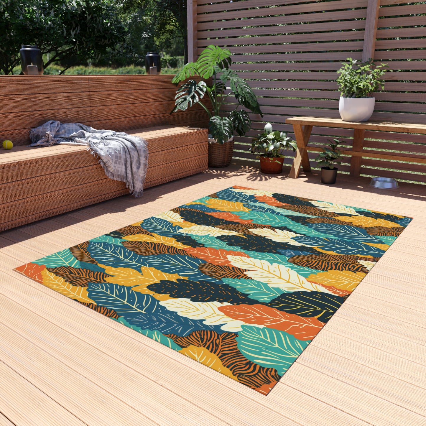 A colorful African-inspired outdoor rug with a bold leaf pattern in yellow, orange, teal, and green lies on a light wooden deck. There’s a wicker bench with a throw blanket, potted plants, a side table, and a wooden privacy screen in the background.