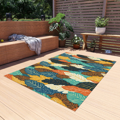 A colorful African-inspired outdoor rug with a bold leaf pattern in yellow, orange, teal, and green lies on a light wooden deck. There’s a wicker bench with a throw blanket, potted plants, a side table, and a wooden privacy screen in the background.