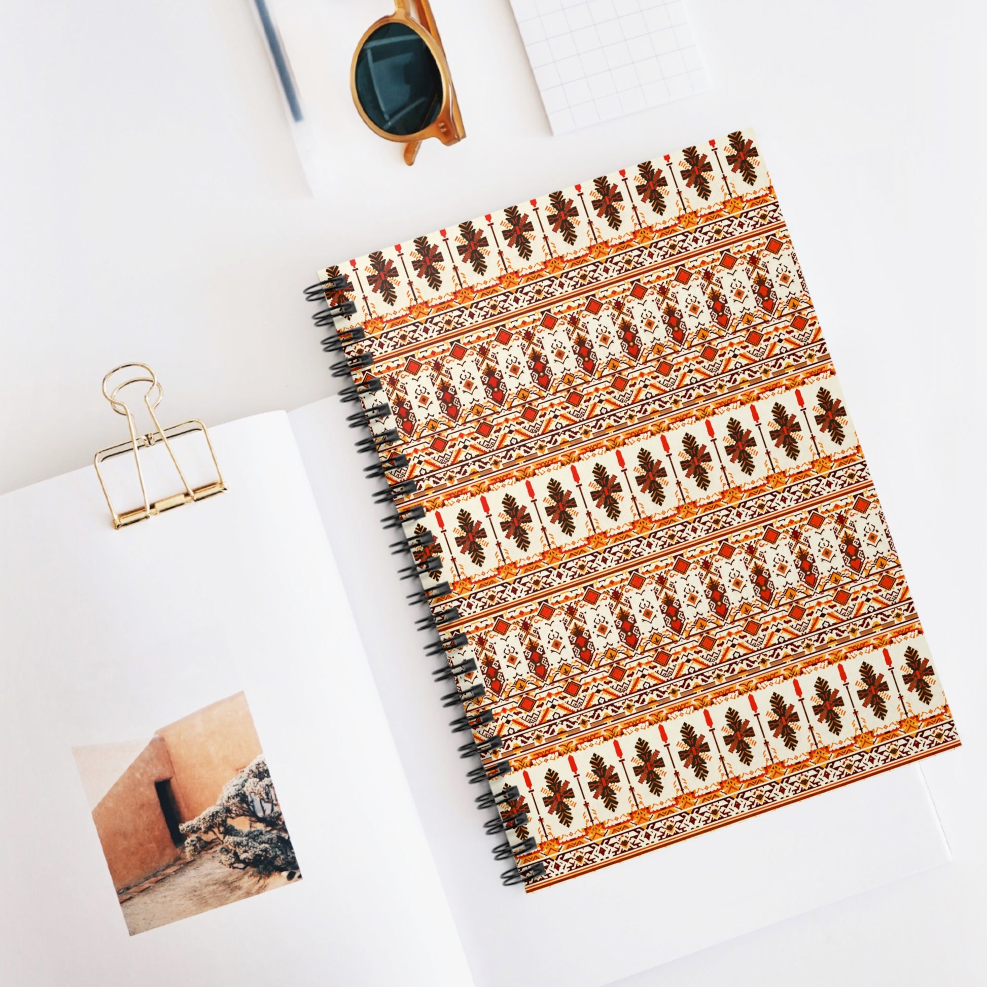 A spiral-bound notebook with African-inspired, red, orange, and beige floral and geometric folk patterns lies on a white desk beside an open book with a photo, a gold paper clip, brown sunglasses, and a small grid notepad.