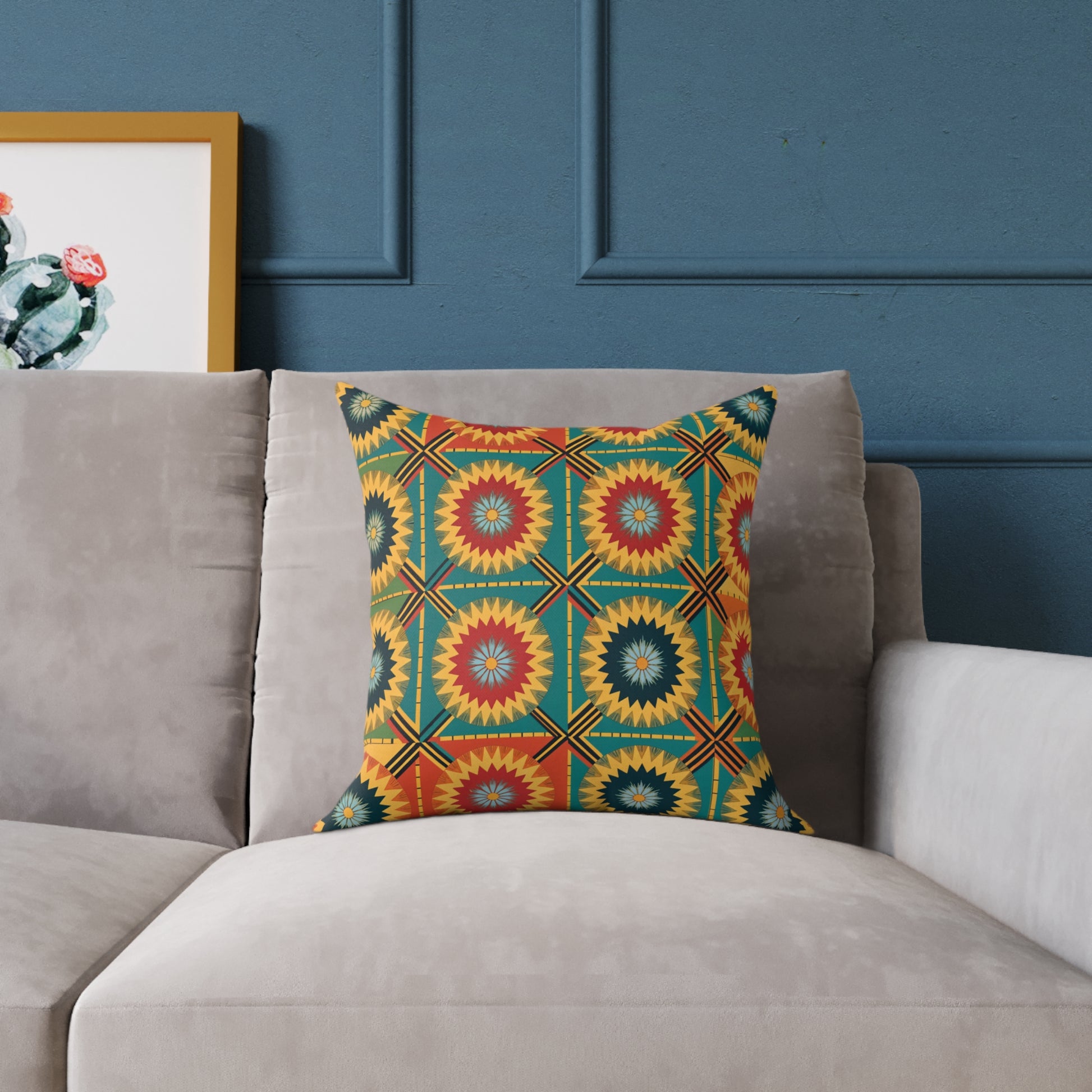A vibrant accent pillow with a bold geometric starburst pattern in red, yellow, green, and blue rests on a light gray sofa. The colorful home decor scene features a blue paneled wall and a framed cactus print visible on the left.