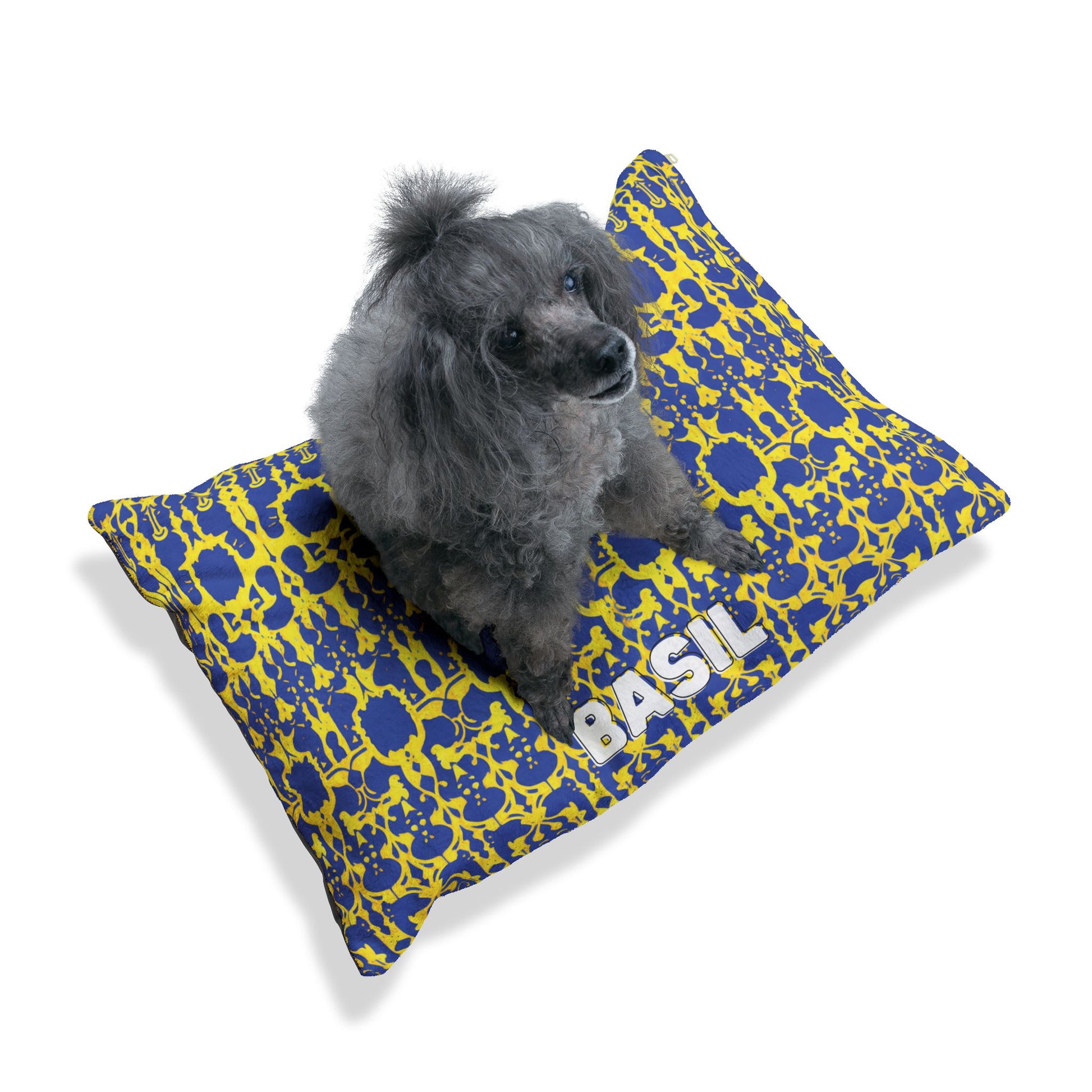 A curly-haired gray poodle sits on a rectangular pillow with a blue and yellow abstract pattern. This personalized pet bed features BASIL in bold white letters, offering luxurious comfort against a plain white background.