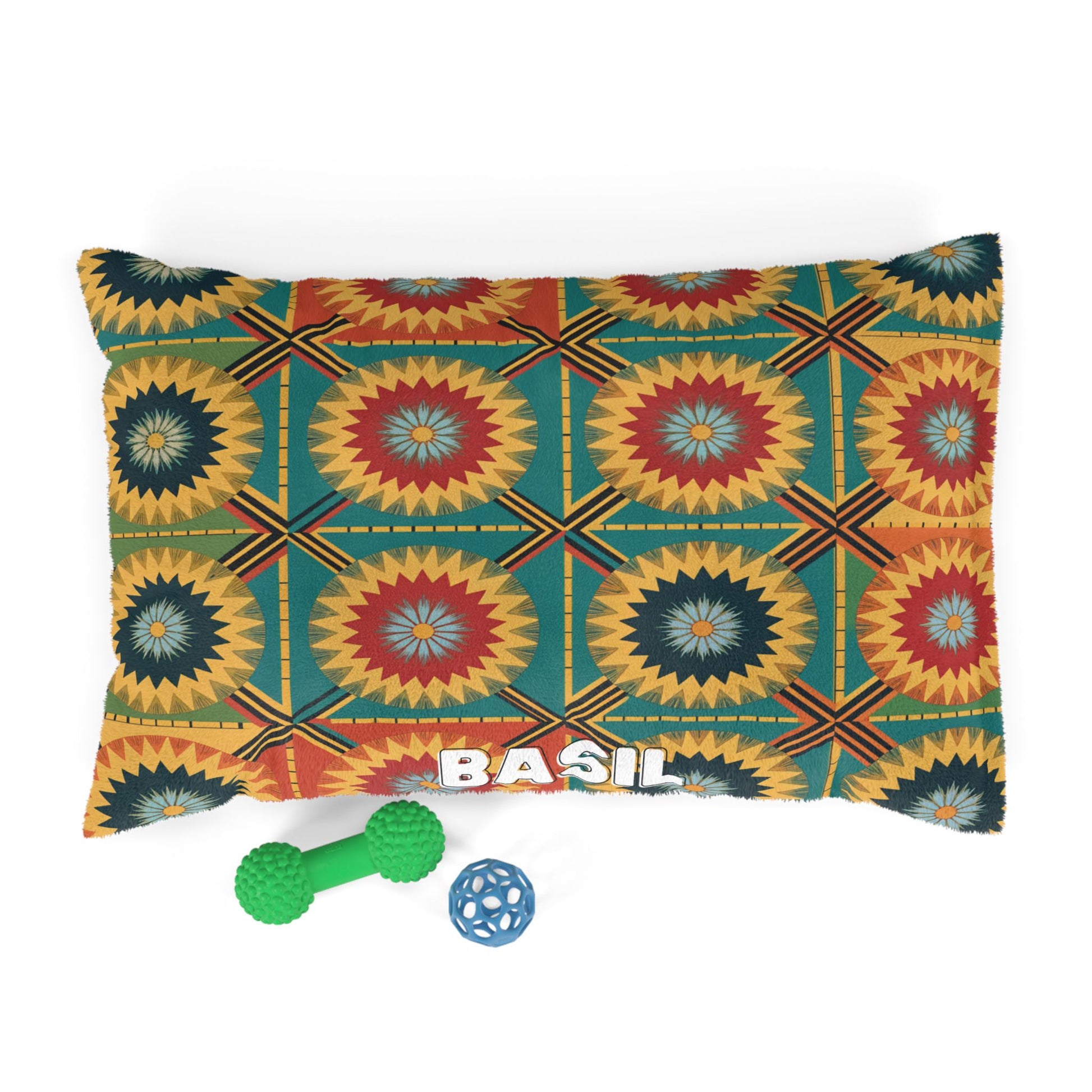 A rectangular pillow crafted from cozy materials features a colorful geometric sunburst pattern and the name BASIL in bold white letters. Two green and blue textured dog toys sit next to this personalized design on a white background.