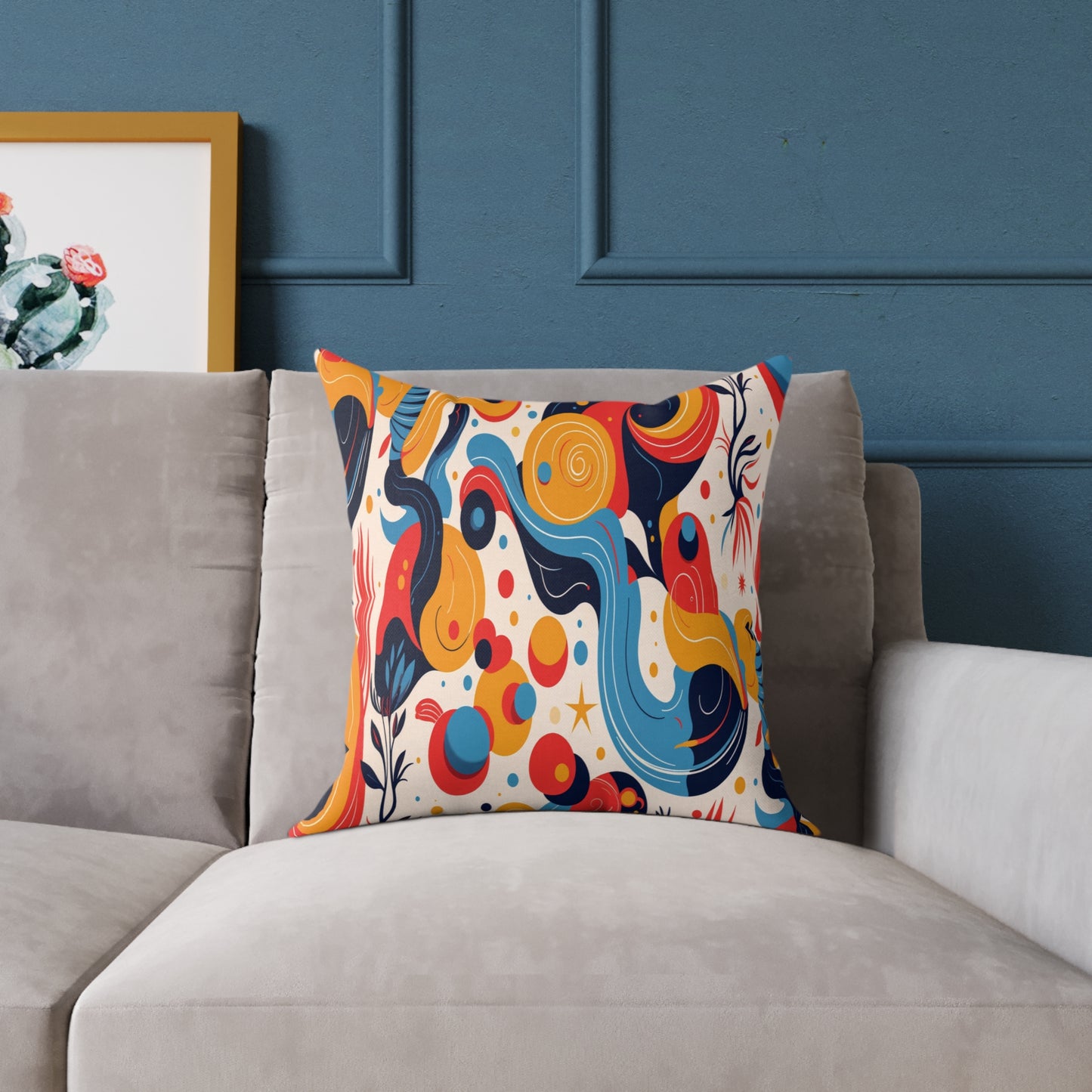 A modern gray sofa with a vibrant, African-inspired pillow featuring abstract swirls, dots, and shapes in blue, yellow, red, and orange. Behind the sofa, a blue wall with molding and a framed cactus artwork is partially visible on the left.