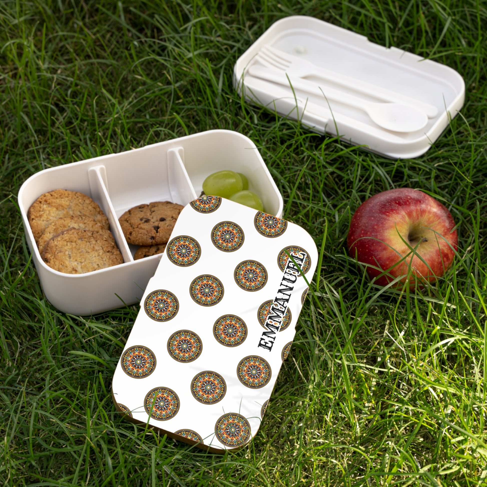 A personalized lunch box with a lid decorated in colorful circular patterns and EMMANUEL in bold letters lies open on grass. Inside this BPA-free lunchbox are cookies and green grapes, with an apple and cutlery nearby.