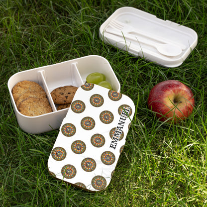 A personalized lunch box with a lid decorated in colorful circular patterns and EMMANUEL in bold letters lies open on grass. Inside this BPA-free lunchbox are cookies and green grapes, with an apple and cutlery nearby.
