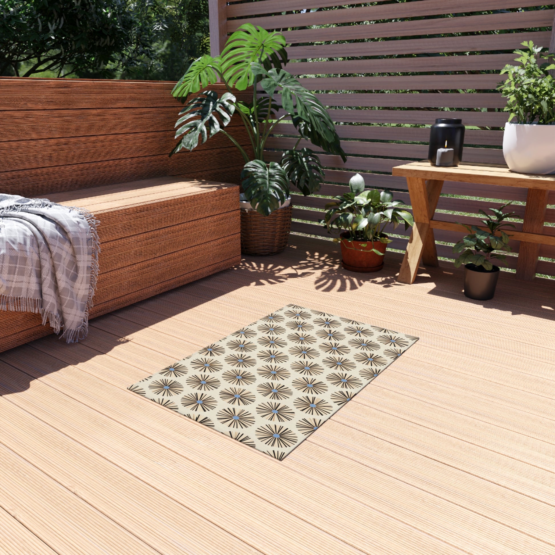 A sunny wooden deck features an African-inspired outdoor rug with geometric patterns, a wooden bench draped in a plaid blanket, potted plants, and a table holding a teapot. Slatted screens provide shade, with greenery visible outside.