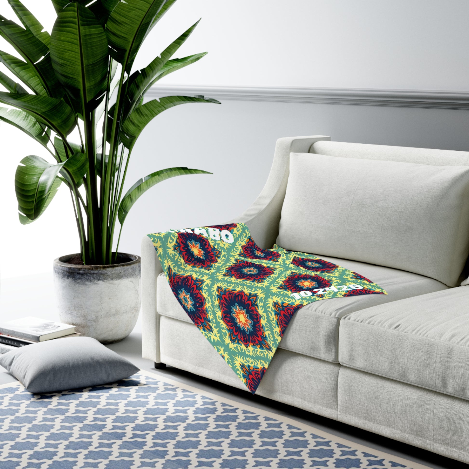 A bright, colorful blanket with an African-inspired geometric pattern is draped over the arm of a modern white sofa. A large potted plant sits nearby, and a blue and white patterned rug covers the floor. A gray pillow rests close by.