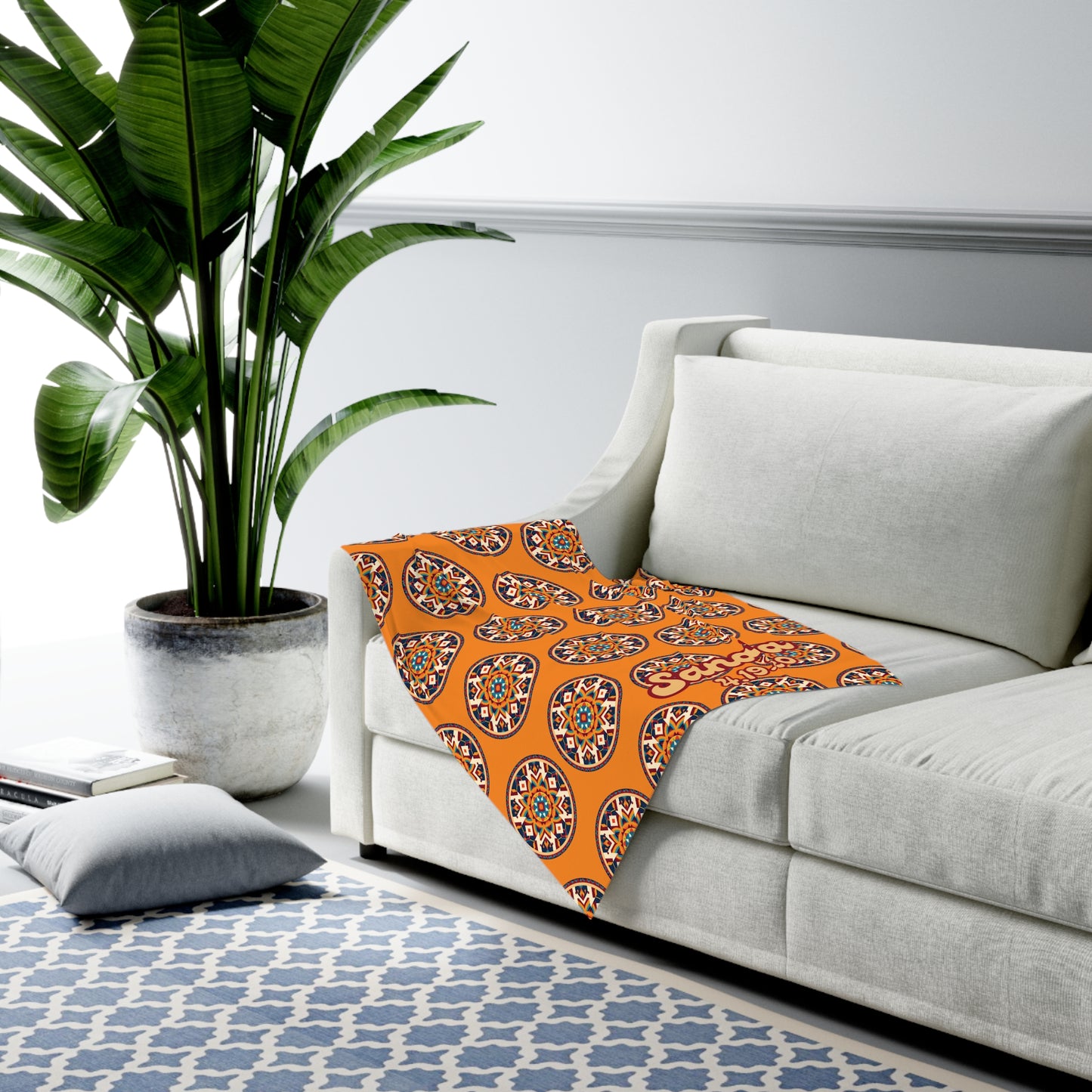 A modern living room with a white sofa, an orange patterned throw draped over the arm, African-inspired decor accents, a large potted plant, a blue and white geometric rug, and a gray floor cushion. The space has light walls and a relaxed, contemporary feel.