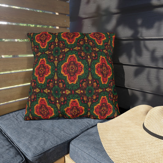 A large square pillow with a bold, intricate African-inspired red and green floral pattern rests on a blue cushioned outdoor bench. Wooden slats and a gray wall set the scene, with a woven straw hat placed nearby in the sunlight.