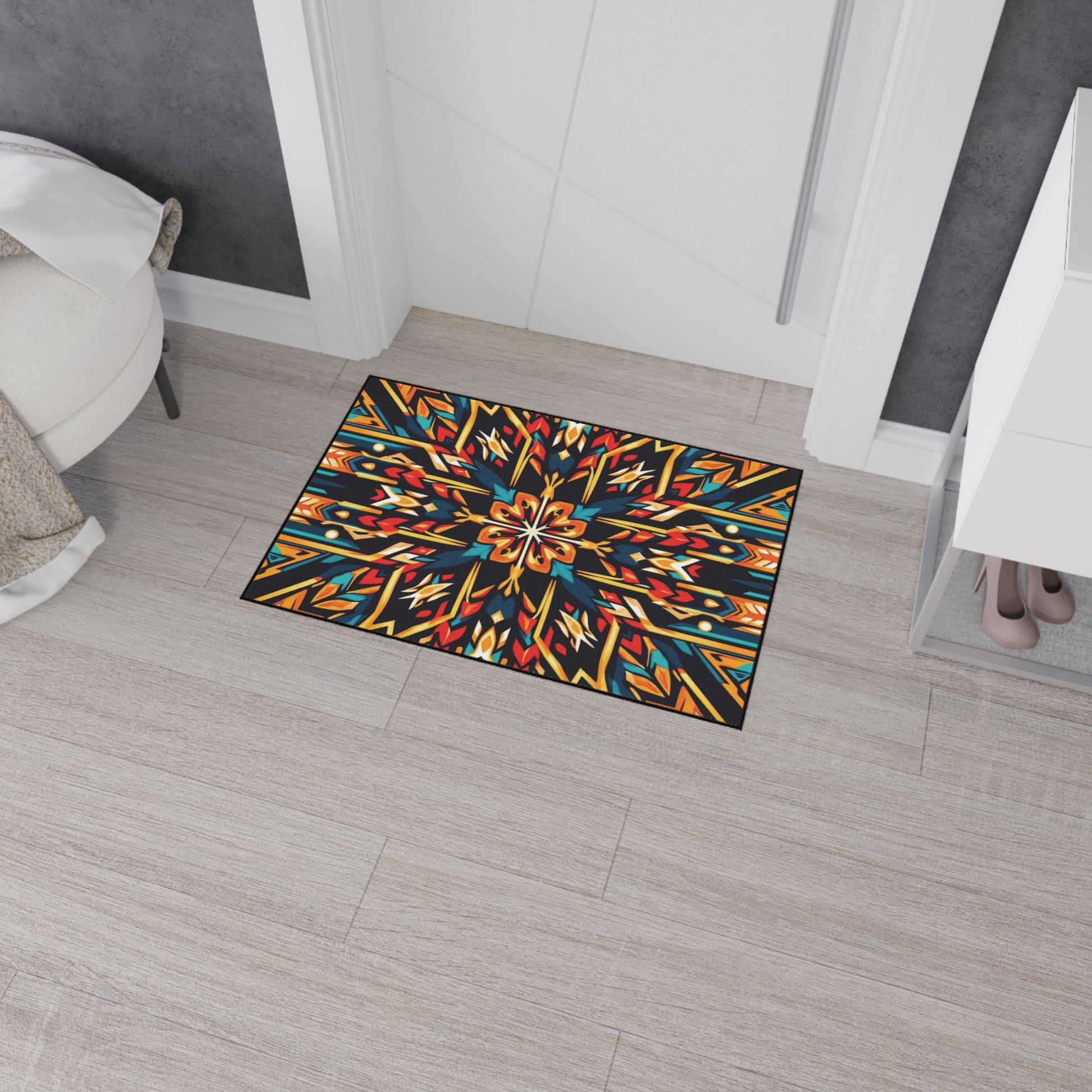 A colorful, African-inspired doormat with a symmetrical, kaleidoscopic geometric pattern in blue, orange, yellow, and red lies on light wood flooring near a white door. The room has a white bench, gray wall, and cabinet with visible shoes inside.