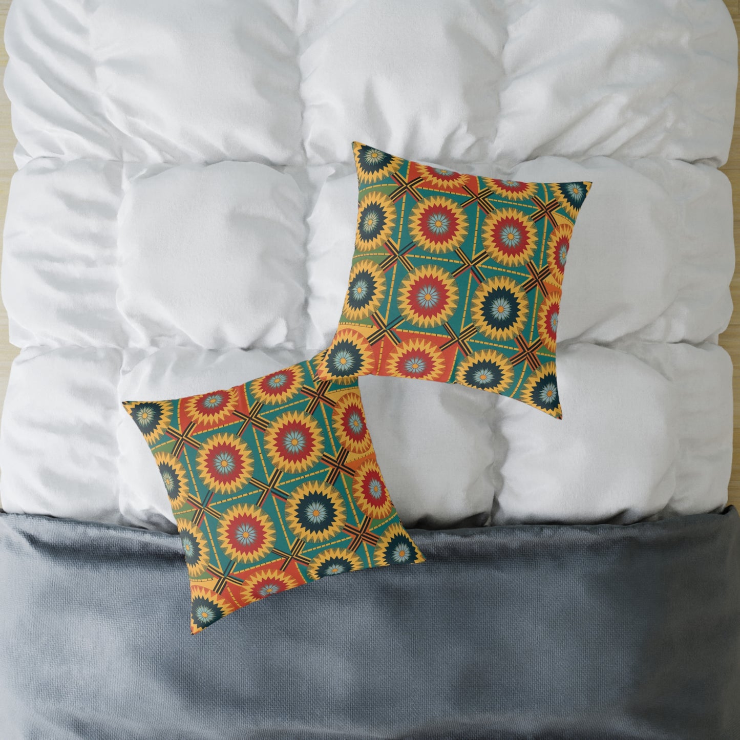 Two decorative pillows with vibrant, geometric starburst patterns in red, yellow, orange, blue, and green serve as colorful accents on a white tufted comforter. A folded gray blanket at the foot completes this inviting home decor scene.