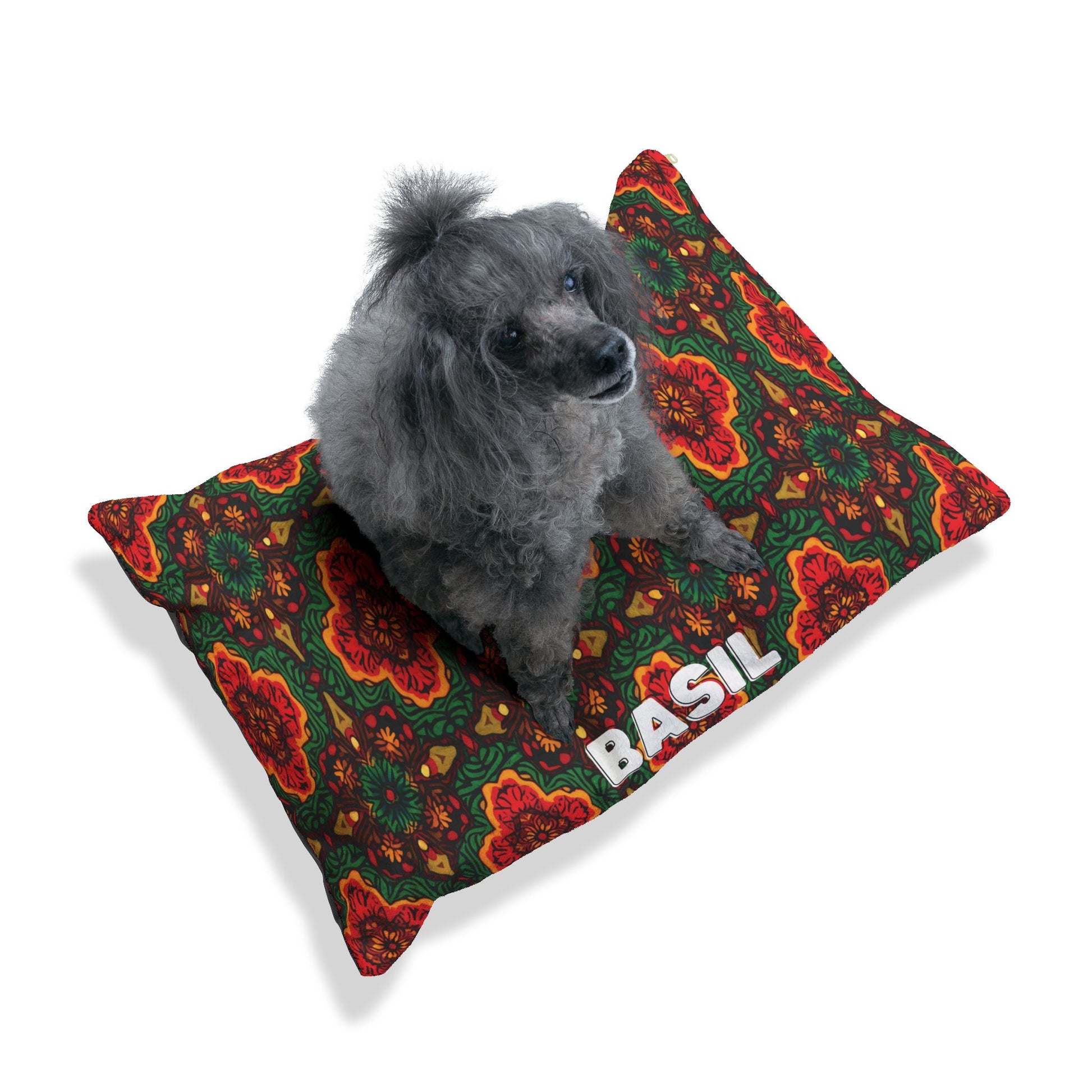 A small gray poodle sits on a personalized pet bed with a colorful, intricate floral pattern in red, green, and orange. The bed offers luxurious comfort and has BASIL written in bold white letters. The background is plain white.