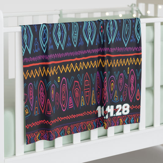 A colorful personalized baby blanket with vibrant geometric and tribal patterns in blue, teal, pink, orange, and yellow drapes over a white crib rail. The blanket features the numbers 11.4.28 in bold white text at the lower right corner. Softly lit background.