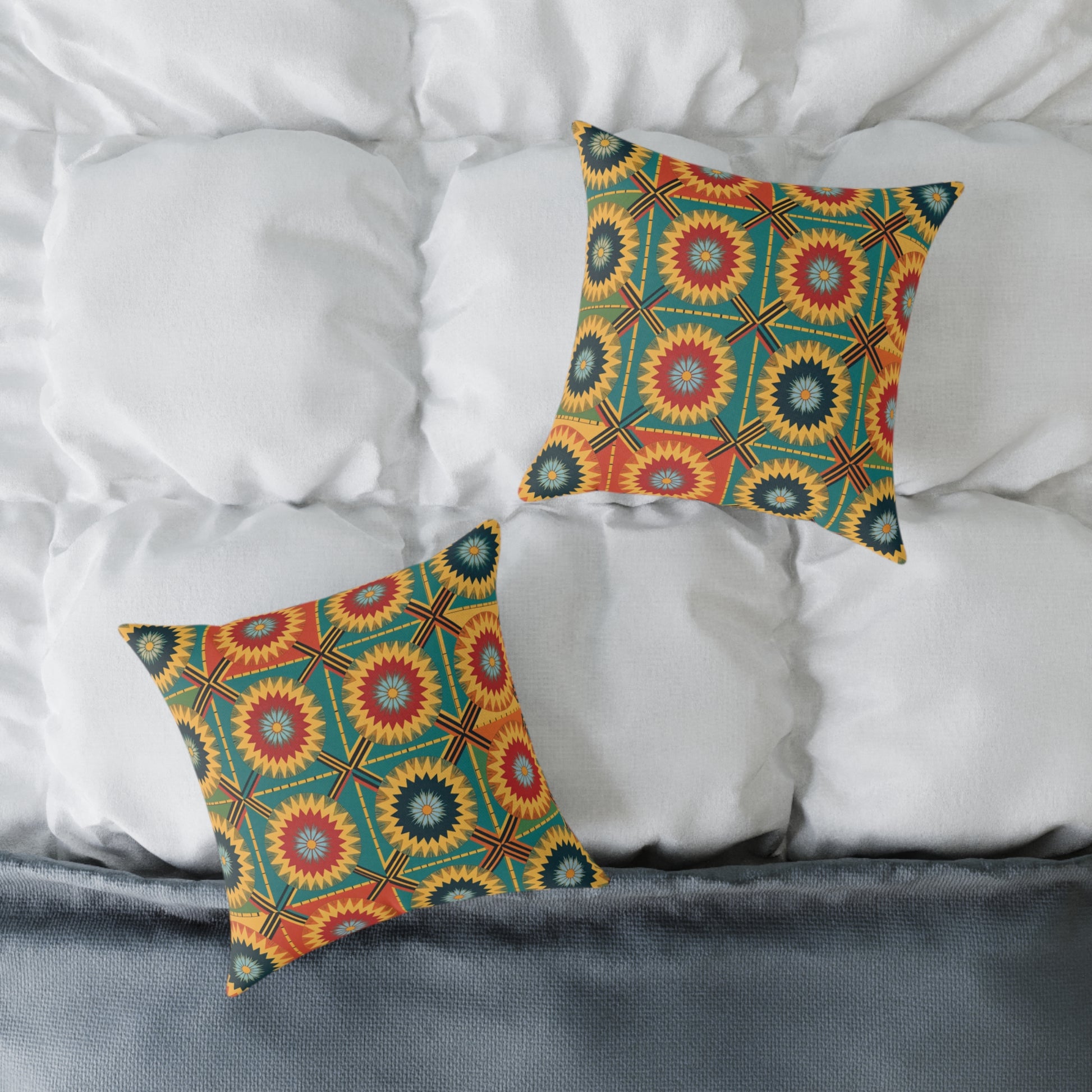 Two square vibrant throw pillows with colorful starburst and geometric patterns in red, yellow, green, and blue rest on a white, tufted bedspread. This colorful home decor is accented by a gray edge or blanket at the foot of the bed.