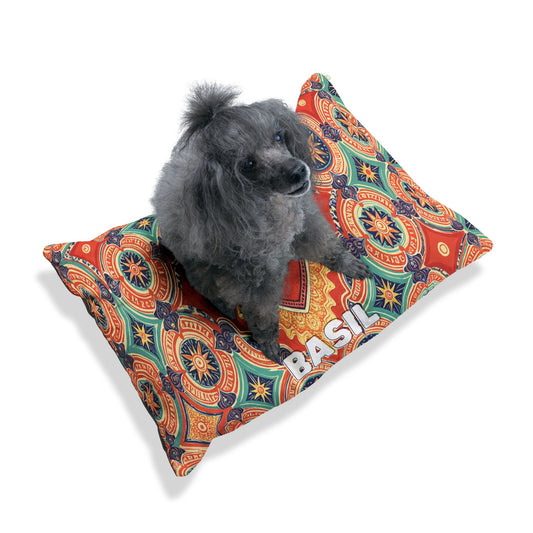 A small gray poodle sits on a stylish, personalized pet bed—a colorful pillow with intricate orange, red, blue, and green designs. The pillow has BASIL printed in bold white letters. The background is white with a soft shadow beneath the bed.
