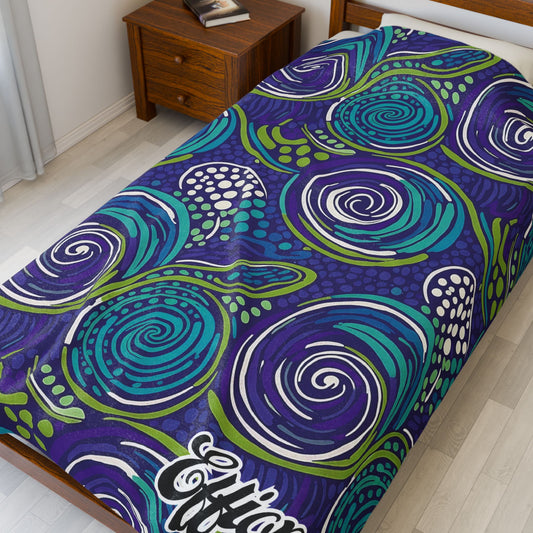 A single bed features a colorful African-inspired blanket with swirling abstract patterns in blue, teal, green, and white. The bed sits beside a wooden nightstand with books, in a bright room with light flooring and white curtains.