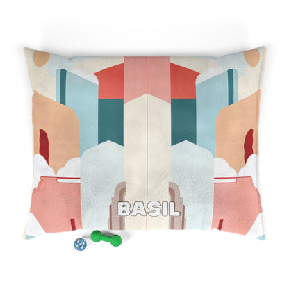 A rectangular dog bed with African-inspired geometric, pastel shapes in tan, teal, coral, and cream. The name “BASIL” is printed in bold white letters at the center. Two dog toys—a blue bone and a green textured ball—rest on the bottom left corner.