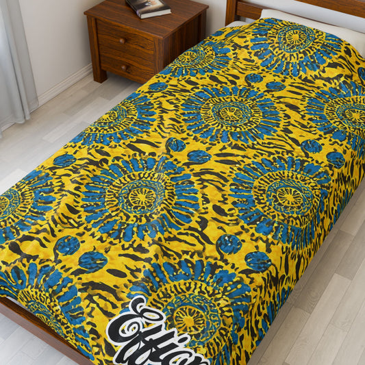 A single bed with a bright yellow and blue African-inspired blanket featuring circular, sunburst-like designs and animal print motifs. The word “Effort” is visible at the bottom corner. A wooden nightstand beside the bed holds books.