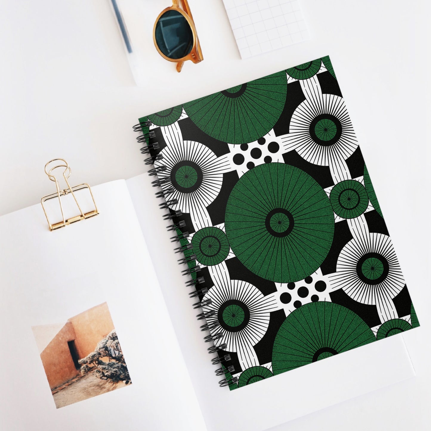 A premium writing experience awaits with this African-inspired spiral notebook, featuring a bold geometric cover in green, black, and white. It sits on a white desk beside an open magazine, gold binder clip, sunglasses, and a gridded notepad.