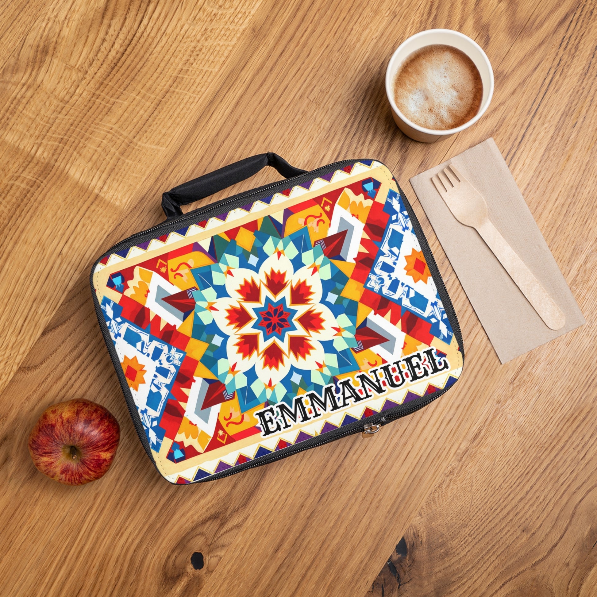 A colorful, African-inspired lunch bag with bold geometric and floral patterns and “EMMANUEL” printed on it sits on a wooden table beside a red apple, a cup of cappuccino, and a napkin with a wooden fork and knife.