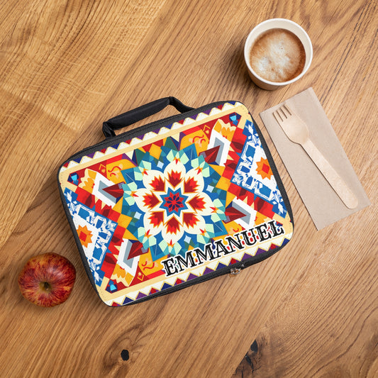 A colorful, African-inspired lunch bag with bold geometric and floral patterns and “EMMANUEL” printed on it sits on a wooden table beside a red apple, a cup of cappuccino, and a napkin with a wooden fork and knife.