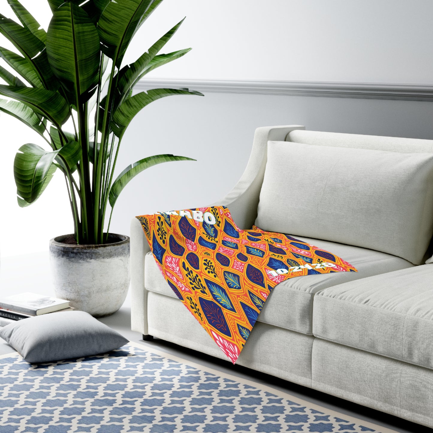 A bright, African-inspired patterned blanket with geometric and floral shapes in yellow, orange, blue, and pink drapes over a light beige sofa. A tall green plant in a white pot sits nearby on the floor. A patterned blue and white rug and a gray cushion are also visible.