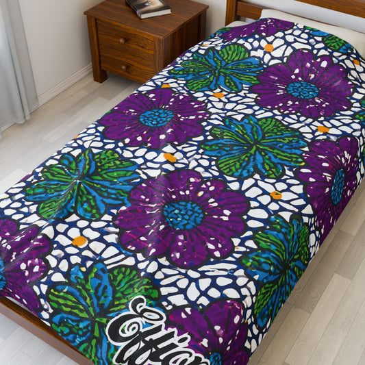 A bed showcases an African-inspired blanket with large purple and green flowers outlined in blue on a white, net-like background. The wooden nightstand holds two closed books, complementing the light floors and white walls.