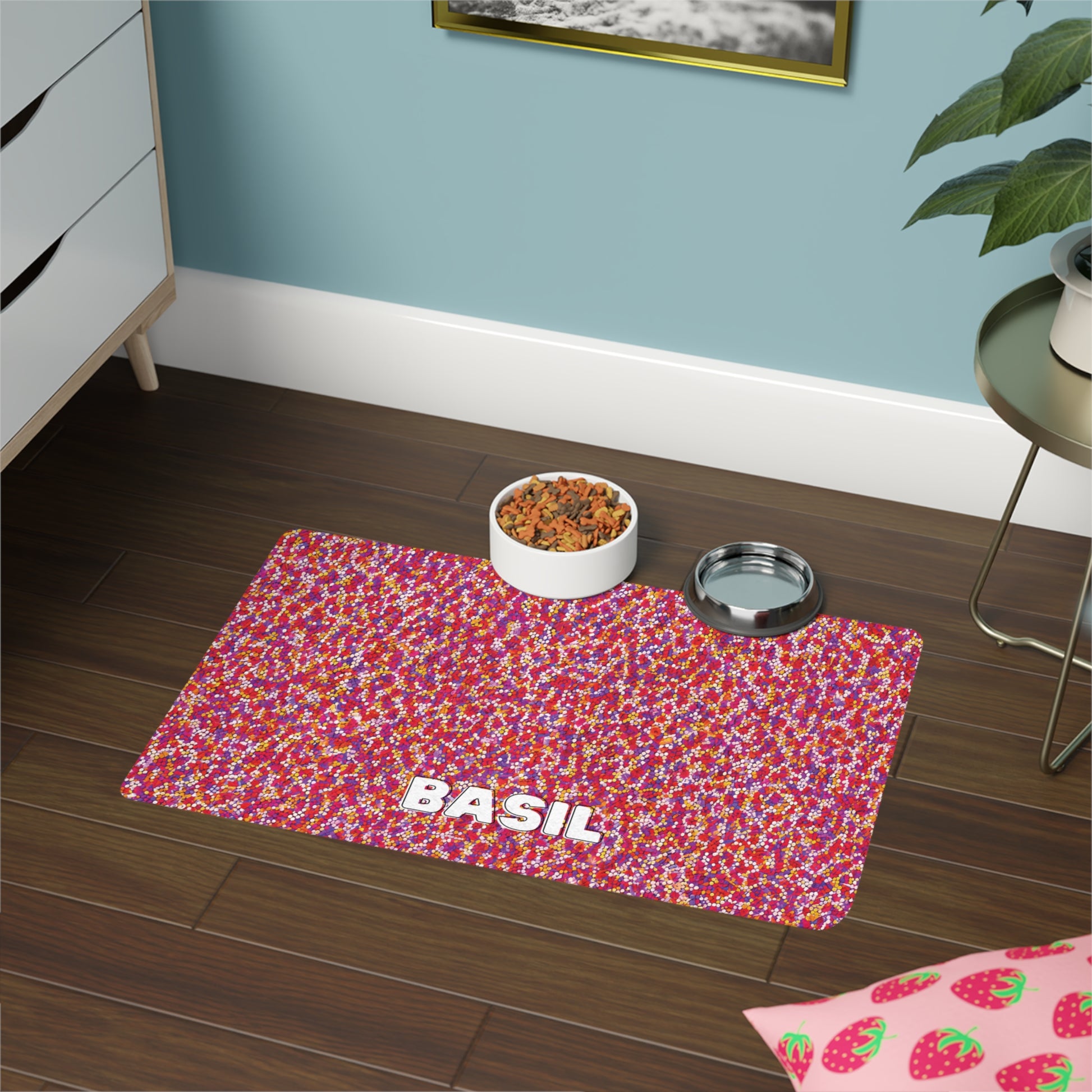 A colorful, African-inspired pet mat with a red, pink, and white confetti-like pattern and the name “BASIL” in bold white letters sits on a dark wooden floor beside a plant, with food and water bowls nearby.
