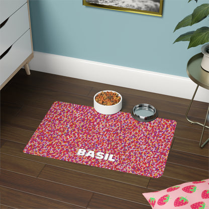 A colorful, African-inspired pet mat with a red, pink, and white confetti-like pattern and the name “BASIL” in bold white letters sits on a dark wooden floor beside a plant, with food and water bowls nearby.