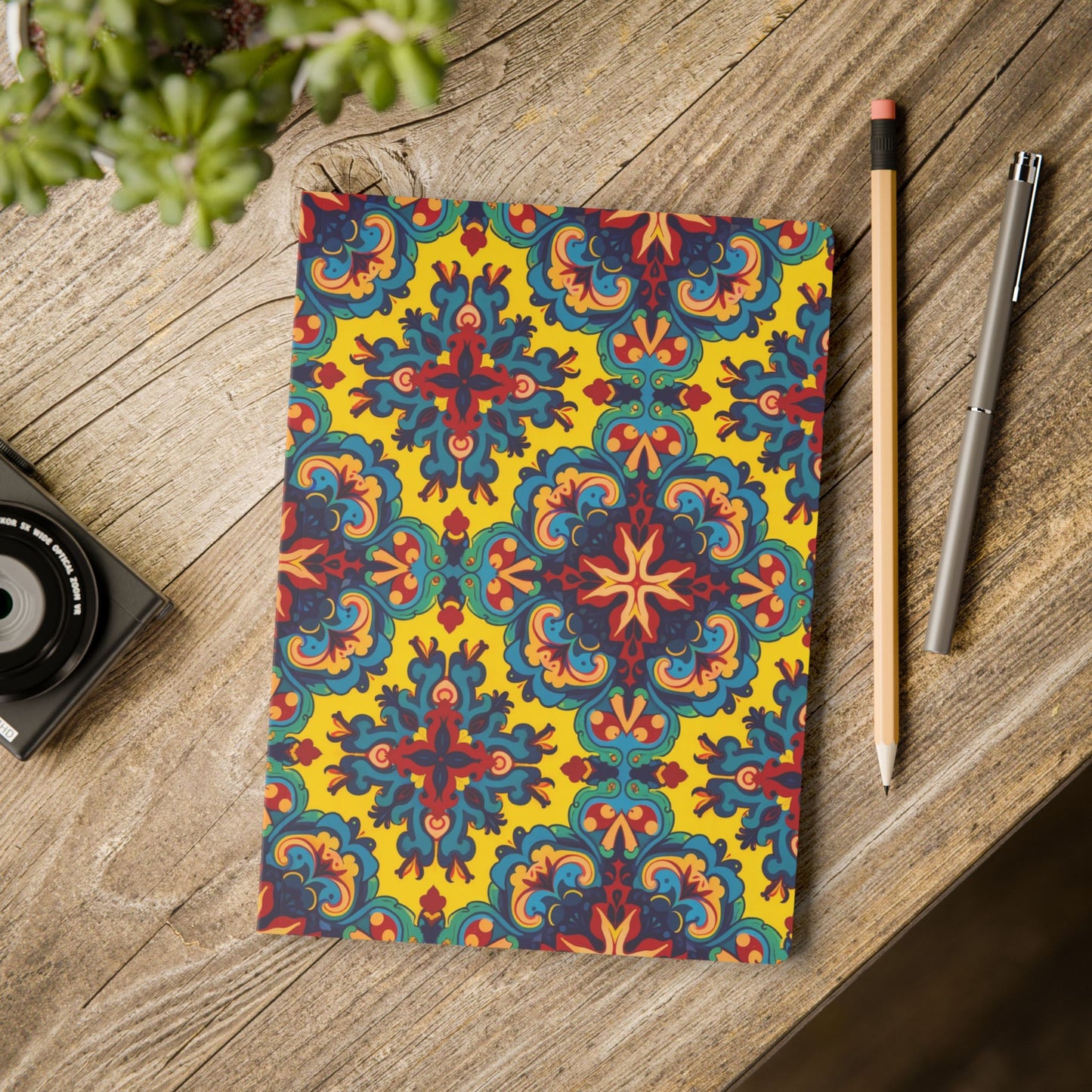 An African-inspired journal with a vibrant, intricate floral and geometric pattern in red, blue, and yellow lies on a rustic wooden table, surrounded by a pencil, pen, camera, and a green succulent, creating a cozy, creative workspace scene.