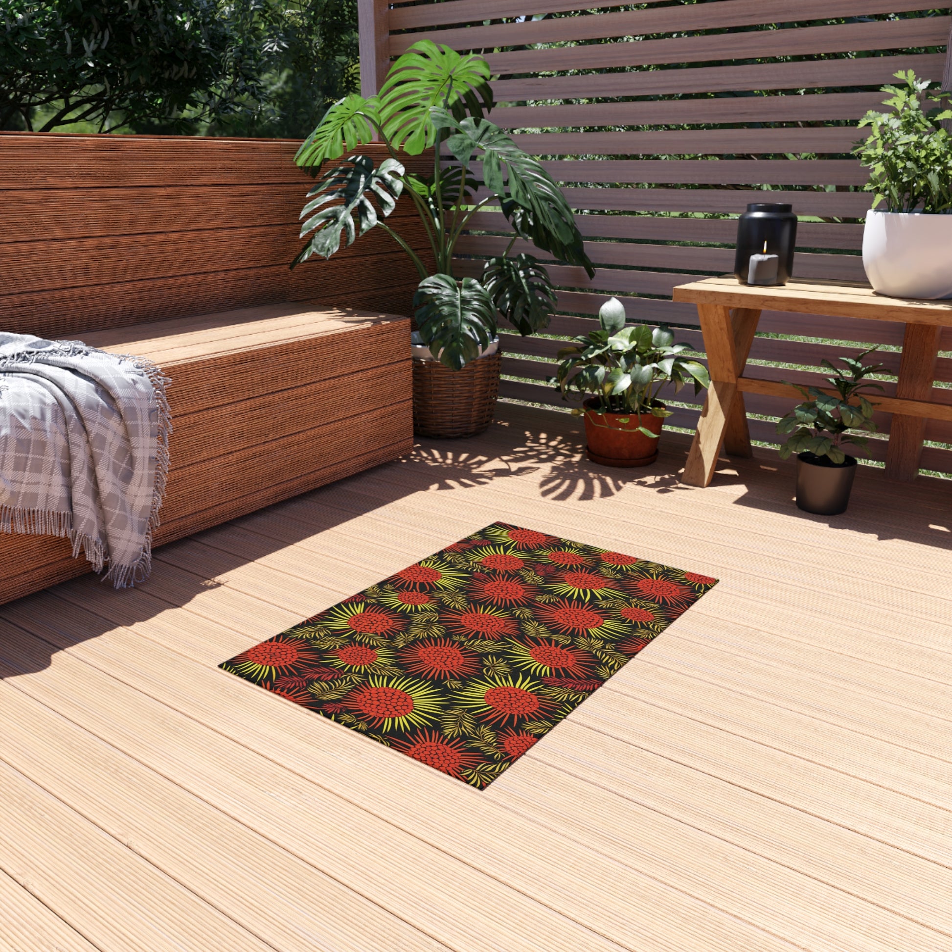 A small rectangular rug with an African-inspired red and green floral pattern lies on a light wooden patio. Nearby are potted plants, a mug, and a wooden bench with a gray blanket. Sunlight filters through slatted fencing and leafy green plants.