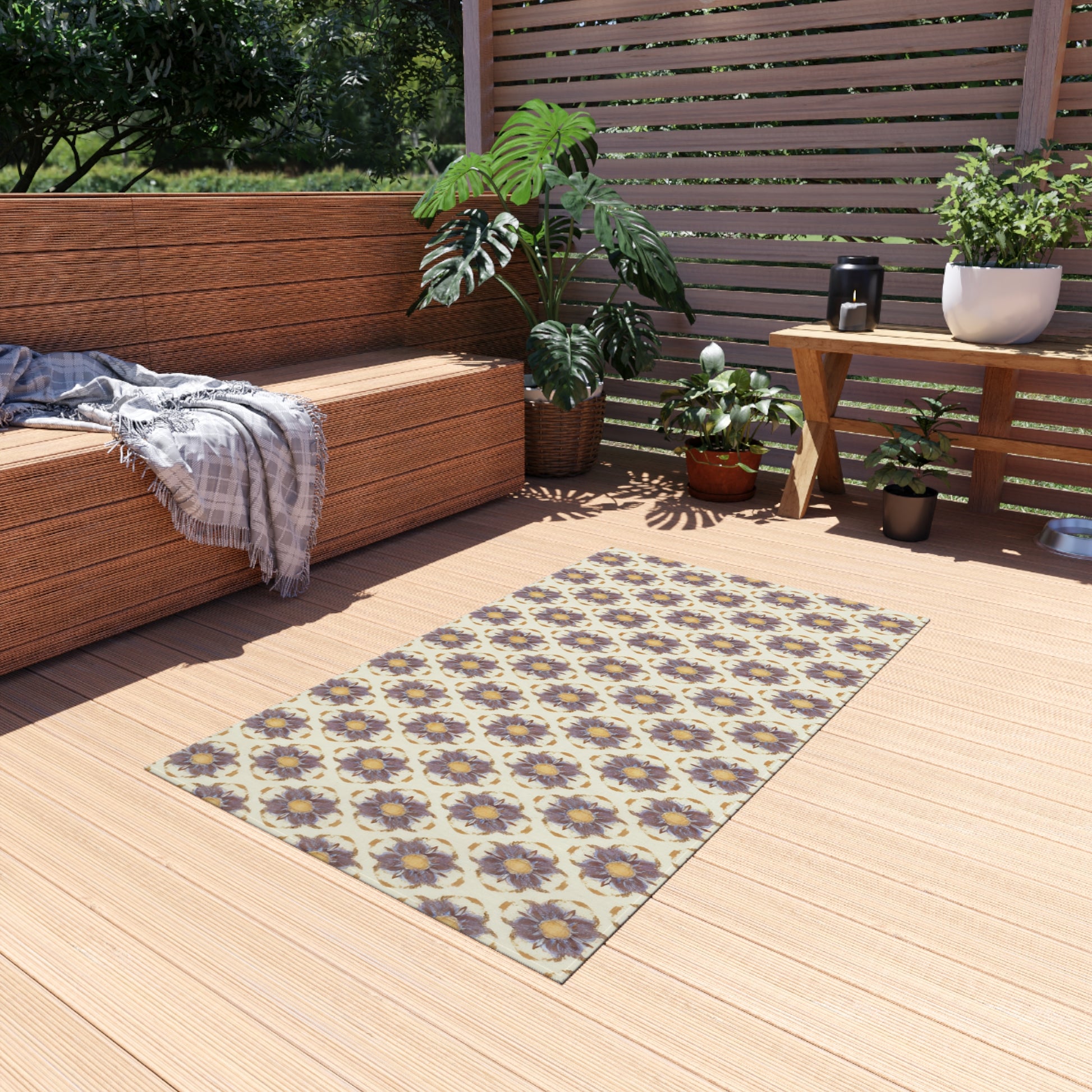 An African-inspired patterned rug with a floral design in beige, purple, and yellow sits on a light wooden deck. Nearby are potted plants, a gray plaid blanket on a wooden bench, and sunlight filtering through slatted fencing.