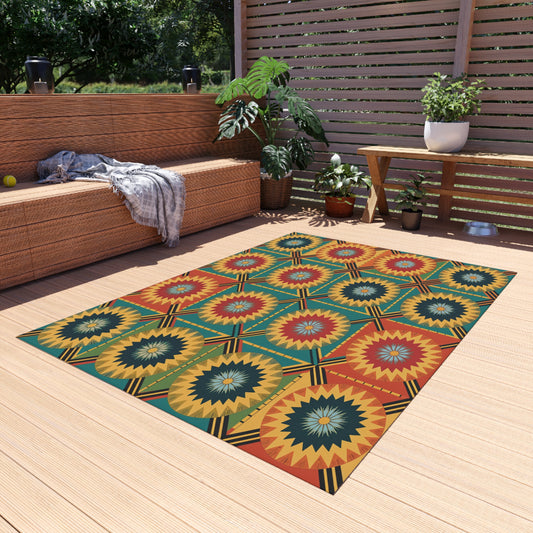 A colorful geometric rug with starburst patterns in red, yellow, teal, and green, inspired by African outdoor rugs and crafted from weather-resistant polyester chenille, lies on a wooden patio near a wicker sofa and potted plants.