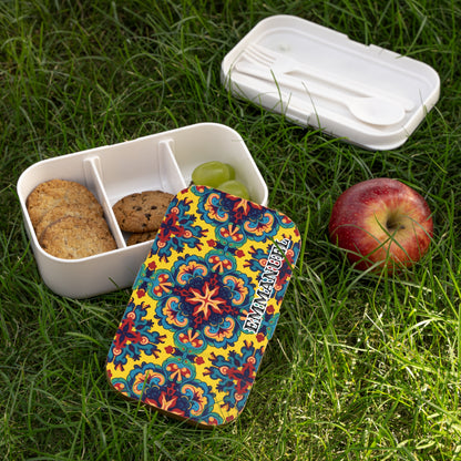 A colorful African-inspired bento lunch box with a vibrant floral pattern sits open on green grass. Inside, there are two cookies, one chocolate chip cookie, and two green grapes. A red apple and white cutlery in a case are beside the lunchbox.